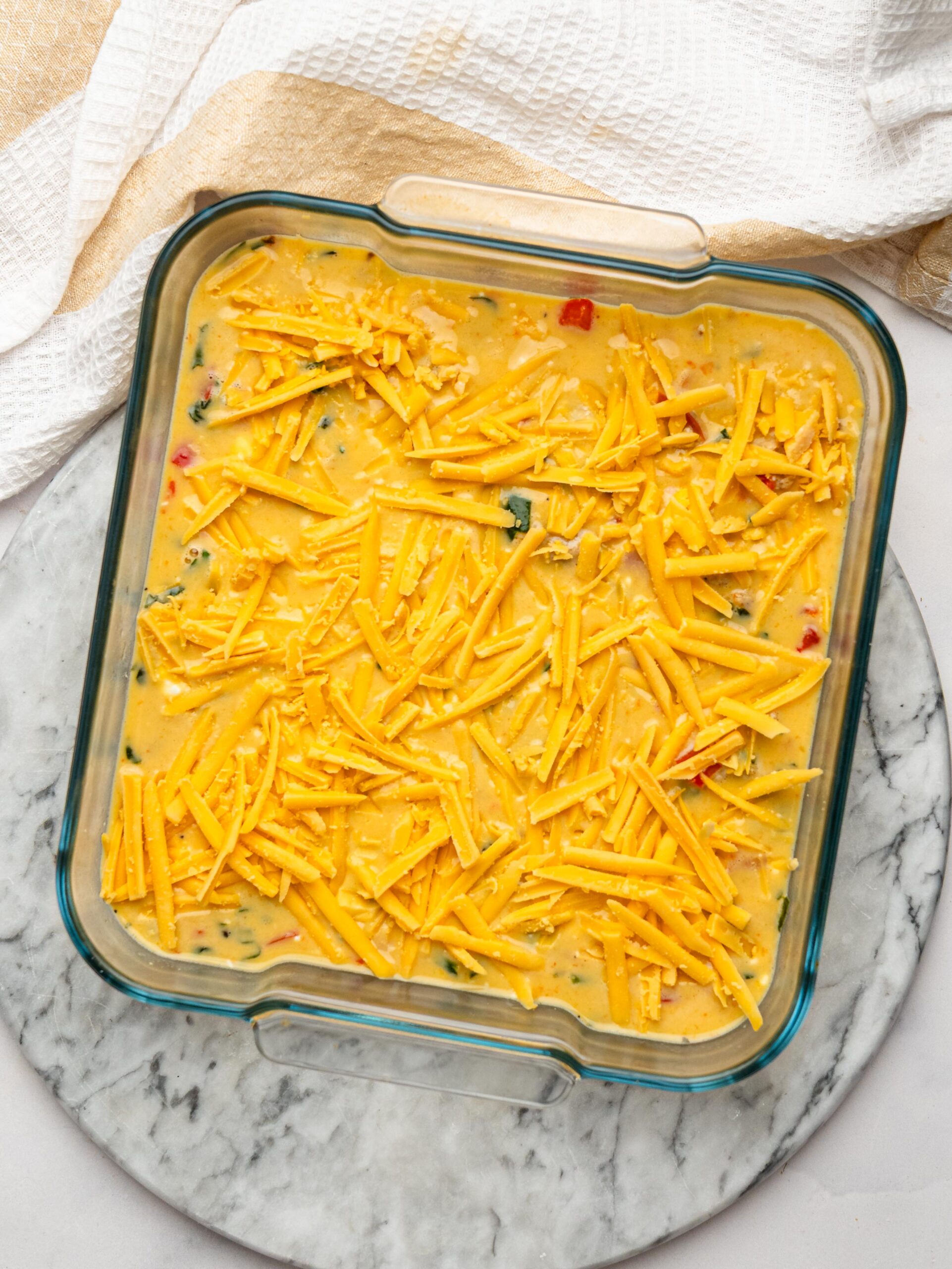 Unbaked egg bake mixture topped with shredded cheddar cheese in a glass baking dish.