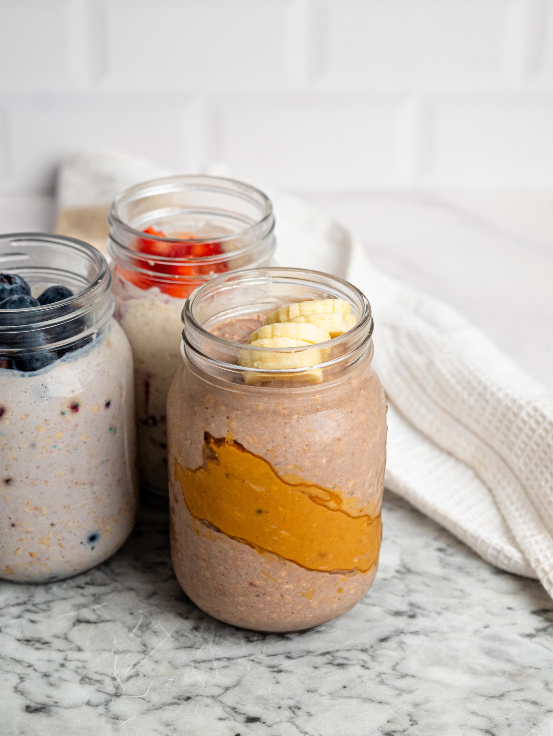 fruit protein overnight oats Three jars of protein overnight oats with banana, strawberries, and blueberries.