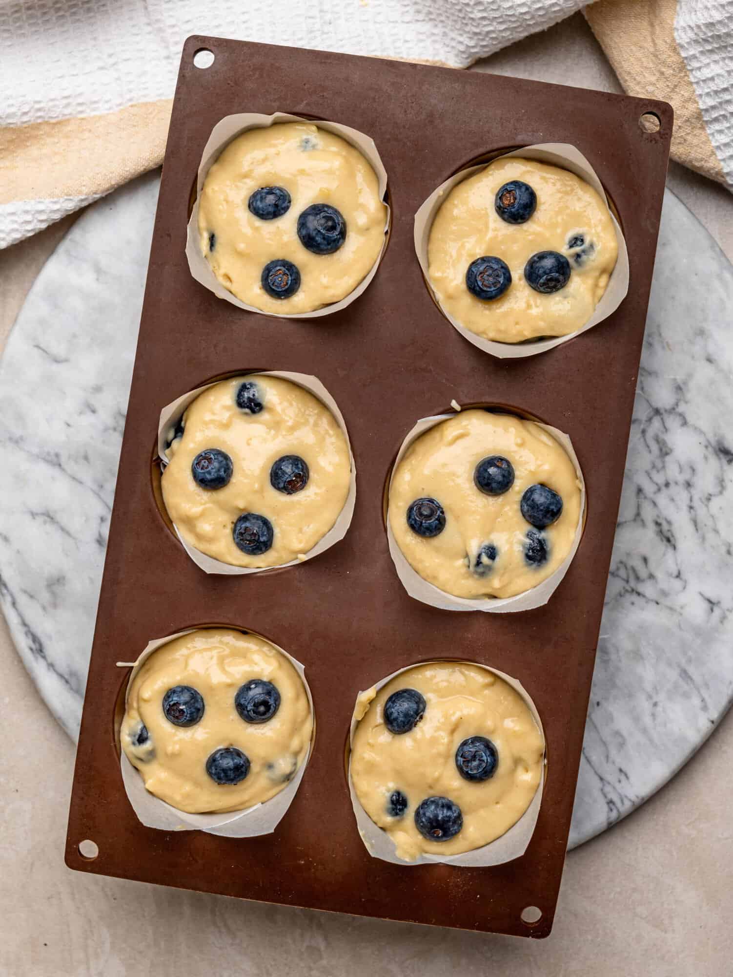 Six blueberry muffins filled with batter and topped with fresh blueberries before baking.