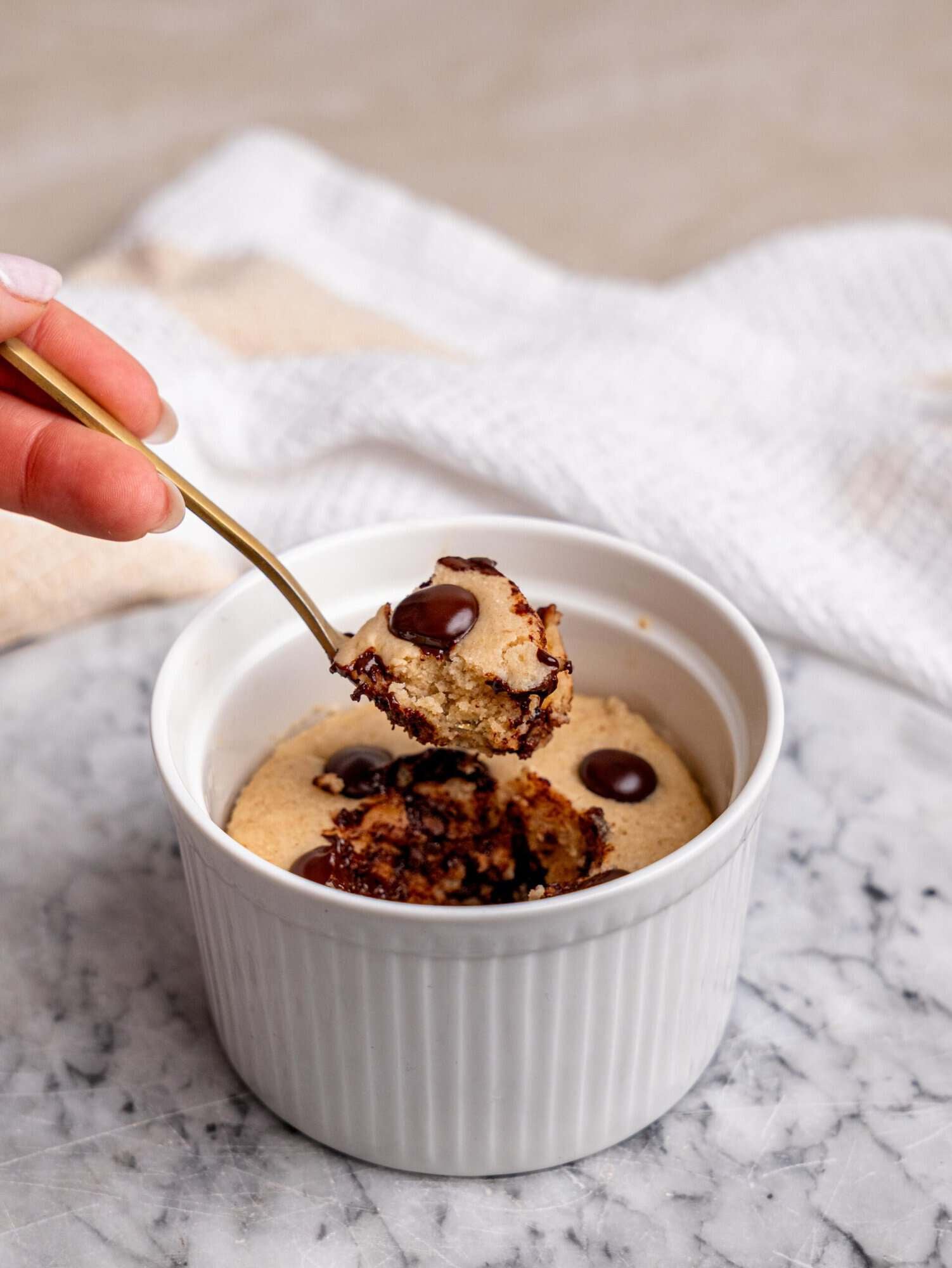 Chocolate chip protein mug cake in a white ramekin, with a spoon lifting a piece of the cake to show its soft, tender texture.
