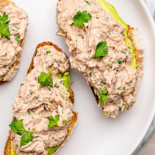 Close-up of creamy tuna salad with chopped pickles and fresh herbs served on toasted bread with avocado.
