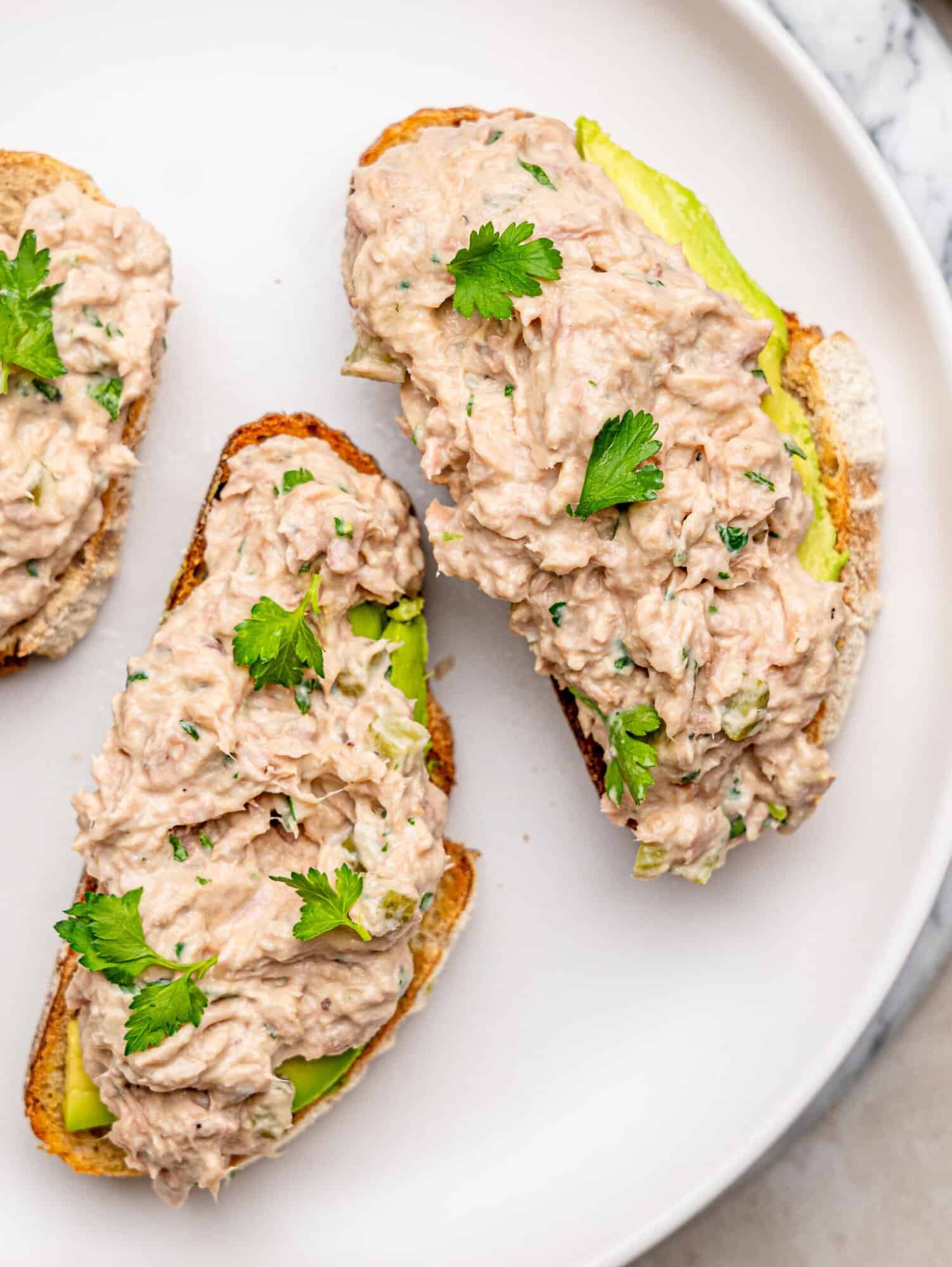Close-up of creamy tuna salad with chopped pickles and fresh herbs served on toasted bread with avocado.