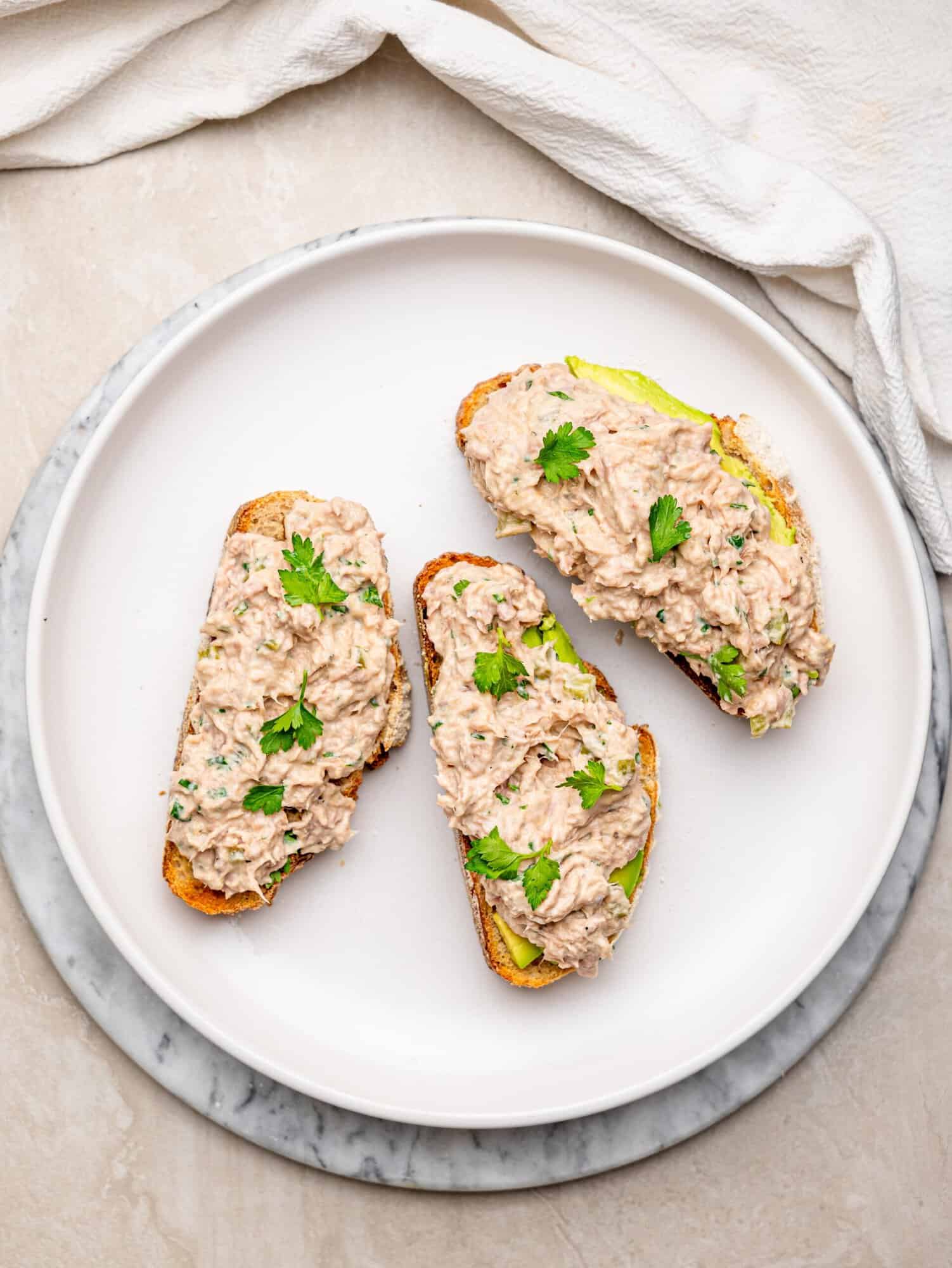 Three slices of toasted bread topped with creamy tuna salad and avocado, garnished with fresh parsley on a white plate.
