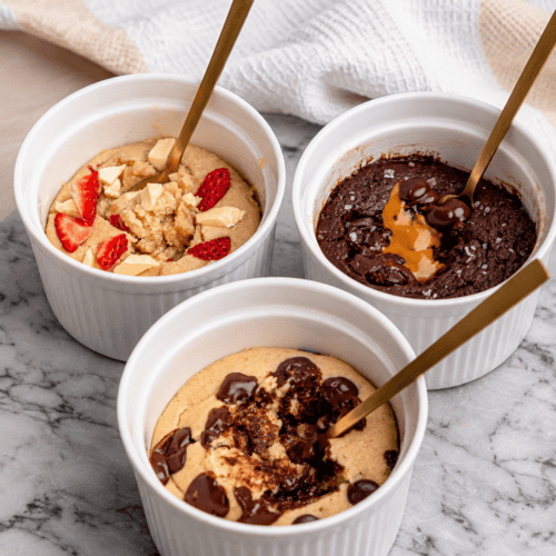 Three protein powder mug cakes in white ramekins, including chocolate chip, Biscoff chocolate, and strawberry white chocolate.