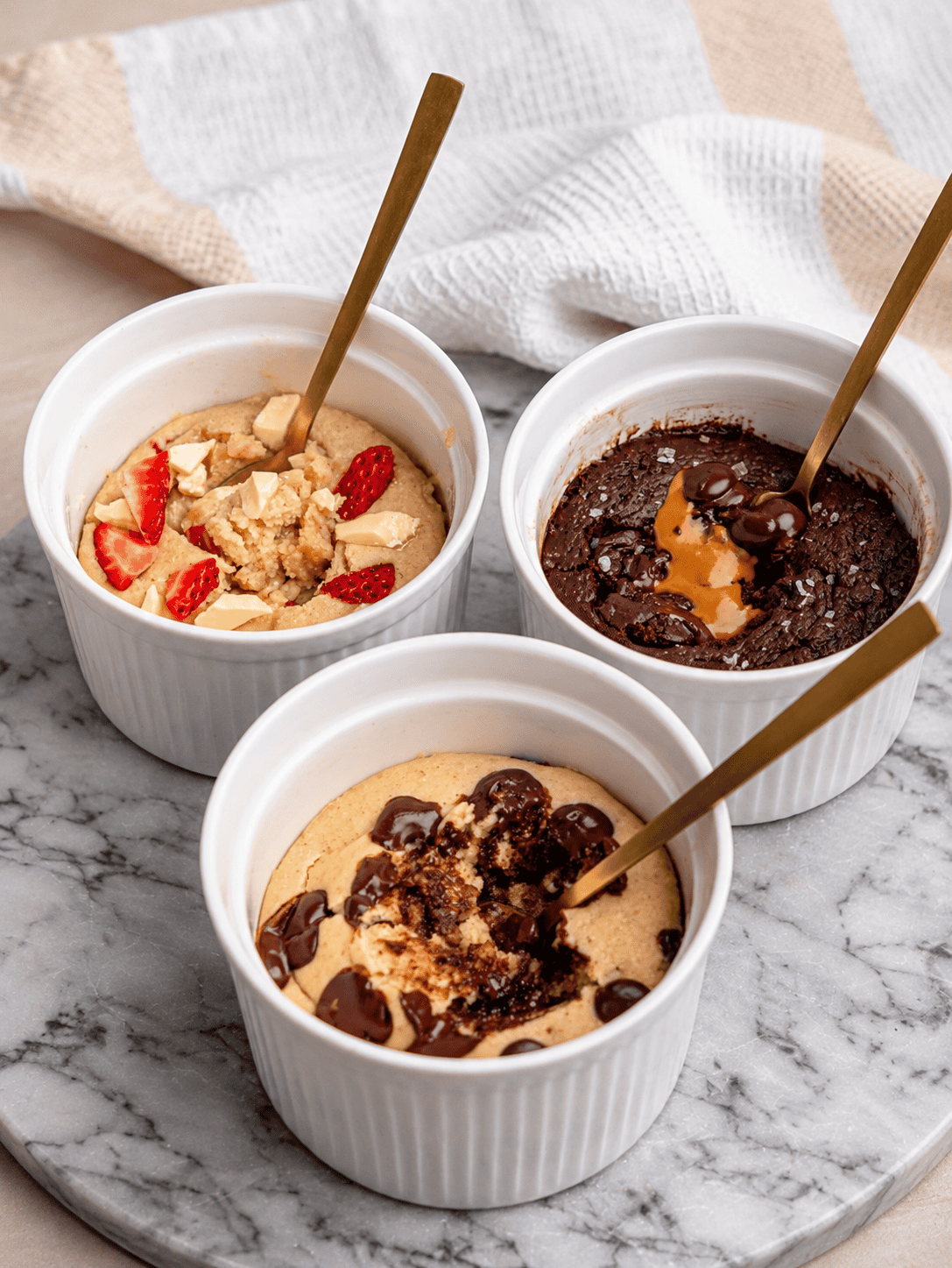 Three protein powder mug cakes in white ramekins, including chocolate chip, Biscoff chocolate, and strawberry white chocolate.