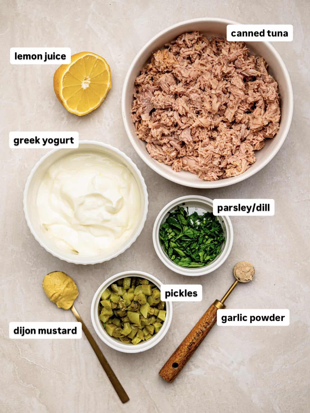 Ingredients for high protein tuna salad arranged overhead with canned tuna, Greek yogurt, lemon, parsley and dill, chopped pickles, Dijon mustard and garlic powder.