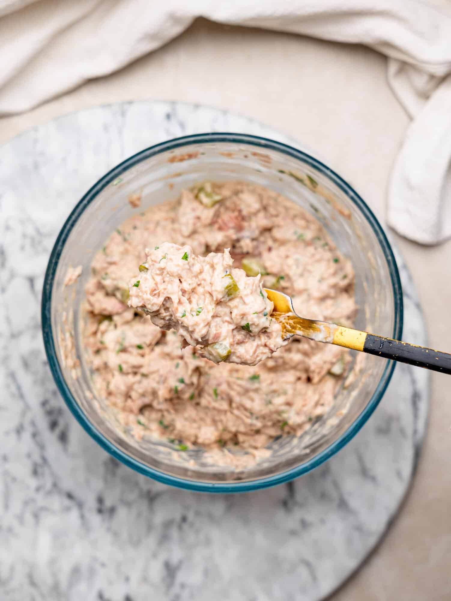 Spoonful of creamy tuna salad with chopped pickles and fresh herbs held over a mixing bowl.