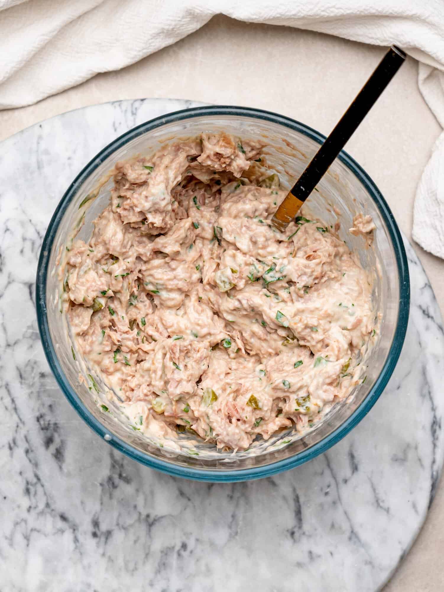 Creamy tuna salad with chopped pickles and fresh herbs in a glass mixing bowl with a spatula.