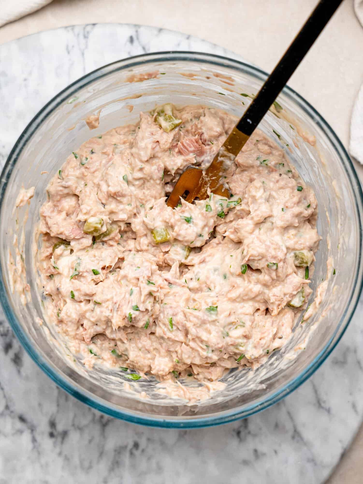 Homemade tuna salad mixed with chopped pickles and fresh herbs in a glass mixing bowl.